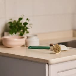 Green and beige silicone back scrubber with a long handle lying on a light countertop with plants in the background. - view 4