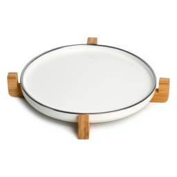 Round white porcelain serving tray with light wood stand on a kitchen counter, holding a small jar and a woven item. - view 3