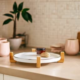 Round white porcelain serving tray with light wood stand on a kitchen counter, holding a small jar and a woven item. - view 1