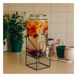 Clear rectangular glass beverage dispenser with a silver spigot and a matching glass lid, sitting on a black wire metal stan… - view 1