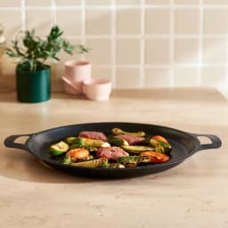 Black round cast iron grill pan with ribbed surface, cooking meat and vegetables on a light kitchen counter. - view 1