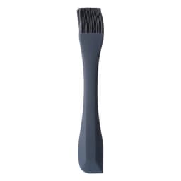 Dark grey silicone basting brush with a long handle lying on a beige kitchen counter next to a small bowl of chopped apples. - view 2
