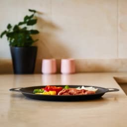 Black round cast iron grill pan with ribbed surface, cooking meat and vegetables on a light kitchen counter. - view 4