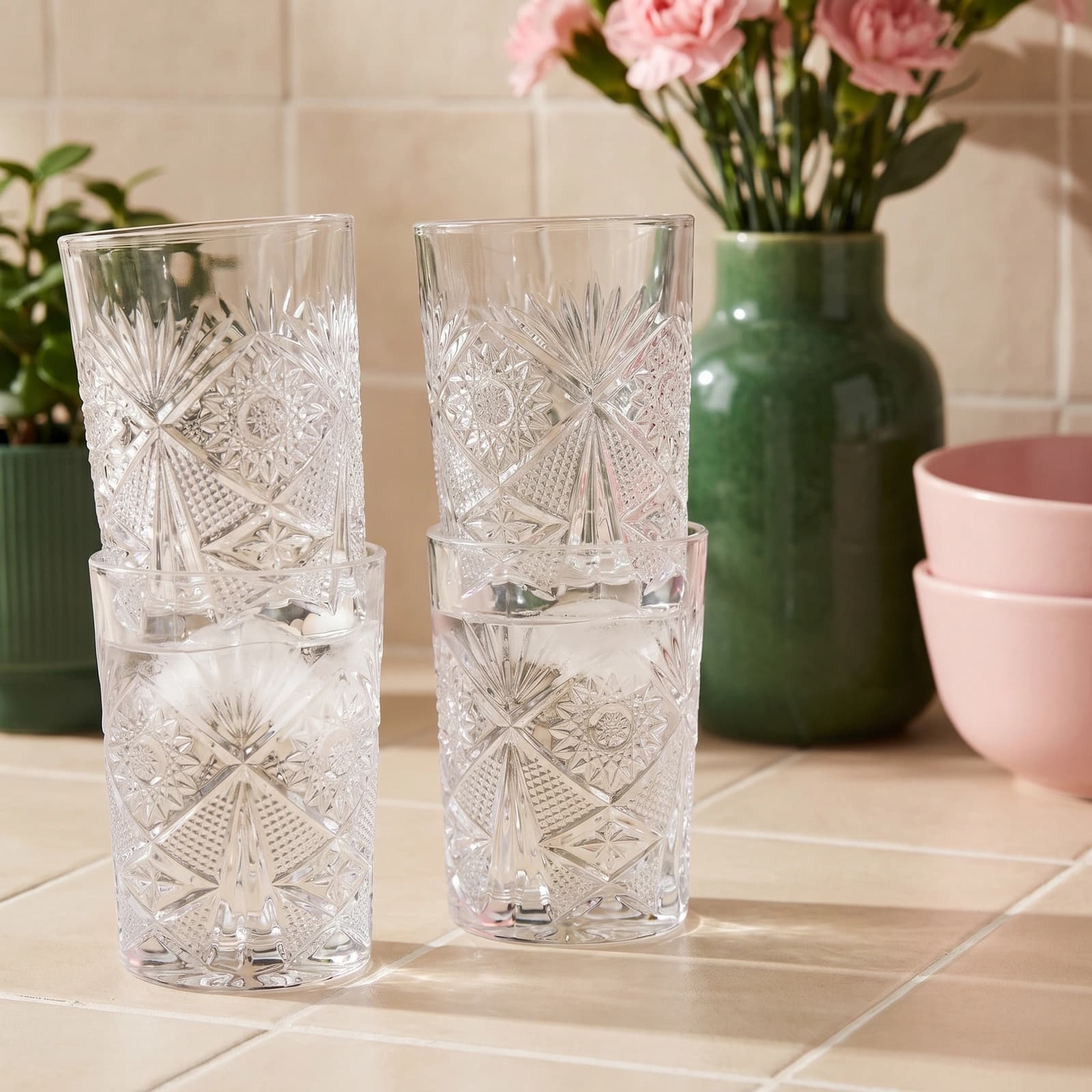 Four clear patterned glass drinking cups, two stacked, on a light tiled surface with plants and pink bowls in background.