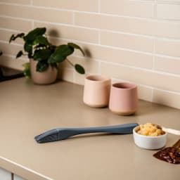 Dark grey silicone basting brush with a long handle lying on a beige kitchen counter next to a small bowl of chopped apples. - view 1