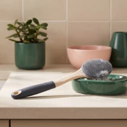 Long-handled silicone bath brush with grey bristles and a beige and dark grey handle, resting on a green soap dish with foam. - view 1
