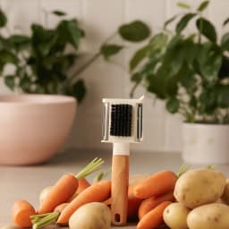 White plastic and wood handle vegetable peeler and scrubber with black bristles, surrounded by fresh carrots and potatoes on… - view 1