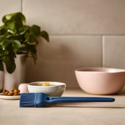 Single dark blue silicone basting brush with a long handle, lying on a light kitchen counter next to small bowls. - view 1
