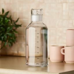 Clear acrylic water bottle with white lid and black time markings on a kitchen counter next to pink mugs. - view 1