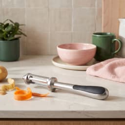 Silver metal vegetable peeler with a black handle peeling an orange carrot on a beige kitchen counter - view 4
