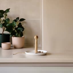 Round white porcelain serving plate with a tall, light wood central handle, on a beige table in a living room setting. - view 6