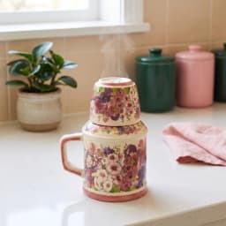 Cream porcelain jug with a handle and a matching cup stacked on top, both adorned with pink and purple floral patterns, on a… - view 1