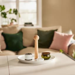 Round white porcelain serving plate with a tall, light wood central handle, on a beige table in a living room setting. - view 3