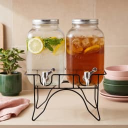Two clear glass beverage dispensers with silver lids and spigots on a black metal stand against a white background. - view 1
