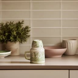 Cream porcelain jug with green floral pattern and matching cup inverted on top, on a kitchen counter. - view 1